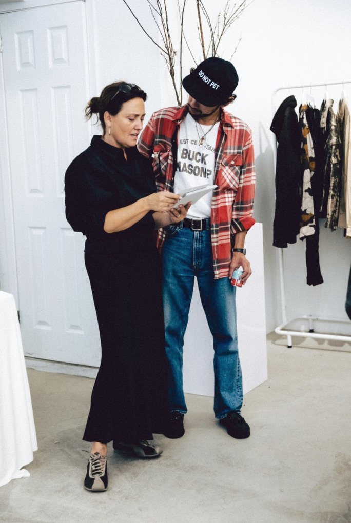 Caroline Rothwell Gerstein reviewing printed materials with a collaborator in a studio, clothing rails behind them.