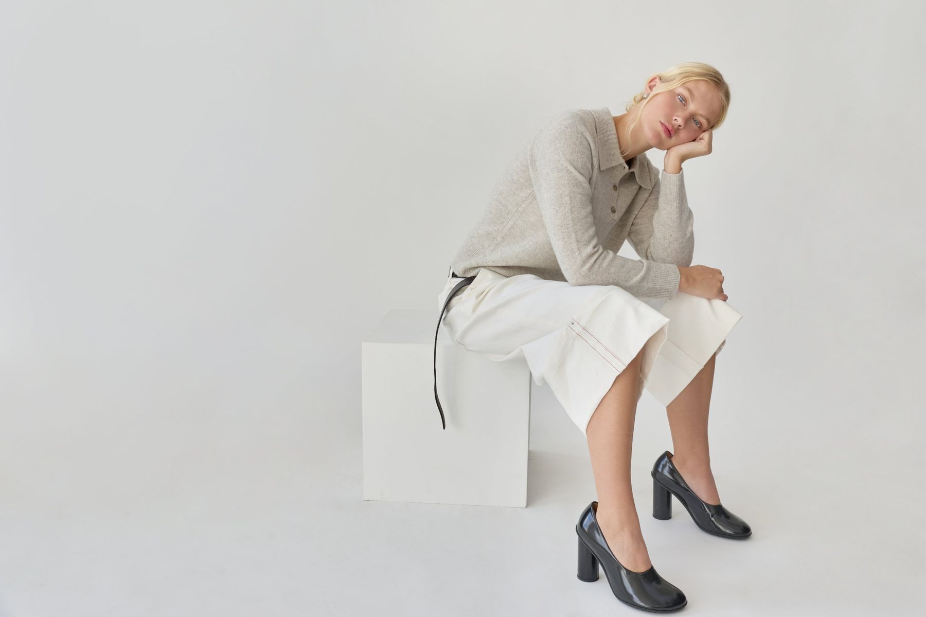A model in a grey knit polo and white denim sits on a white plinth in a pale studio, chin resting on her hand. Fade Out Loud by Honest Thread, Lookbook FW26.