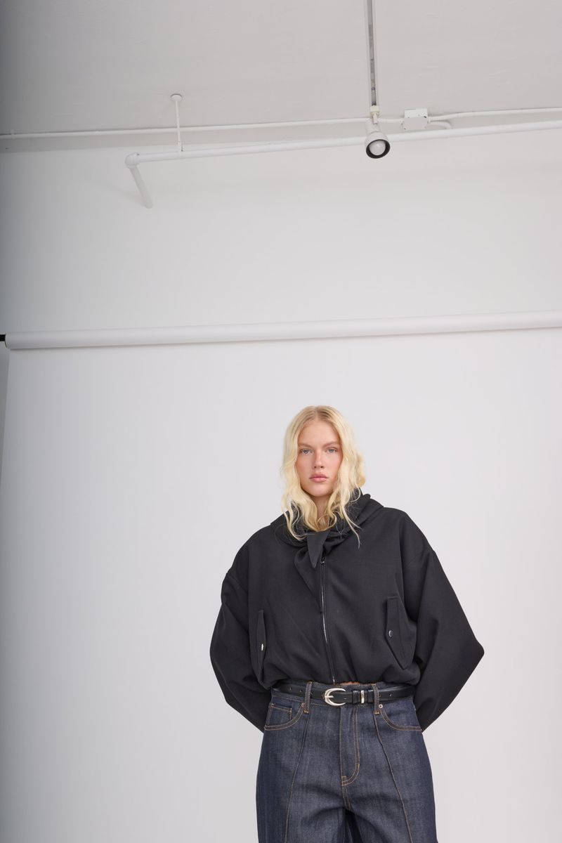 A model in an oversized black jacket and raw indigo denim stands in a white studio under track lights. Fade Out Loud by Honest Thread, Lookbook FW26.