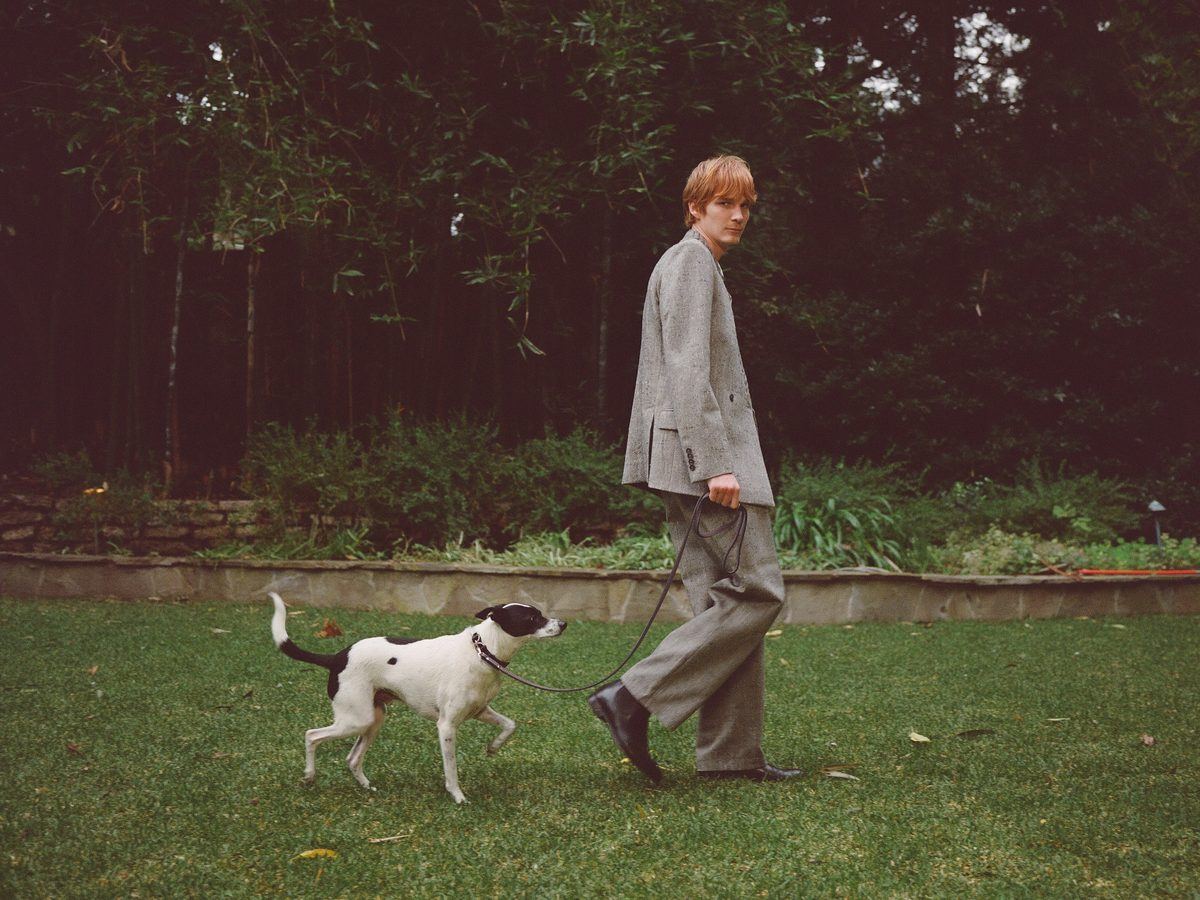 A young man in a tailored grey suit walks a black-and-white dog across a garden lawn. Grayson Kelly Studio, Lookbook FW26.